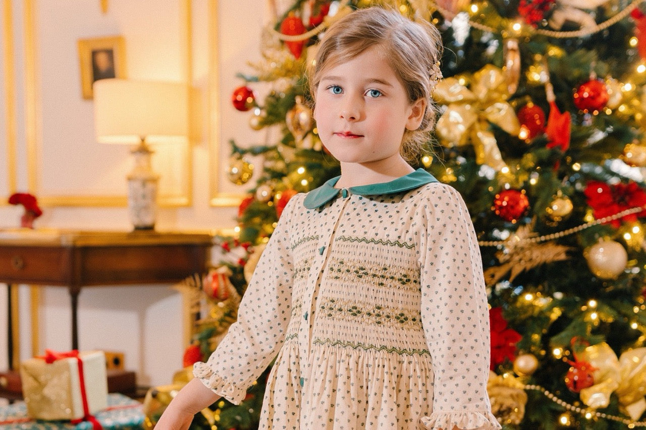 Little girl in a cream and green smocked Christmas dress by Antoinette Paris, standing in front of a decorated Christmas tree