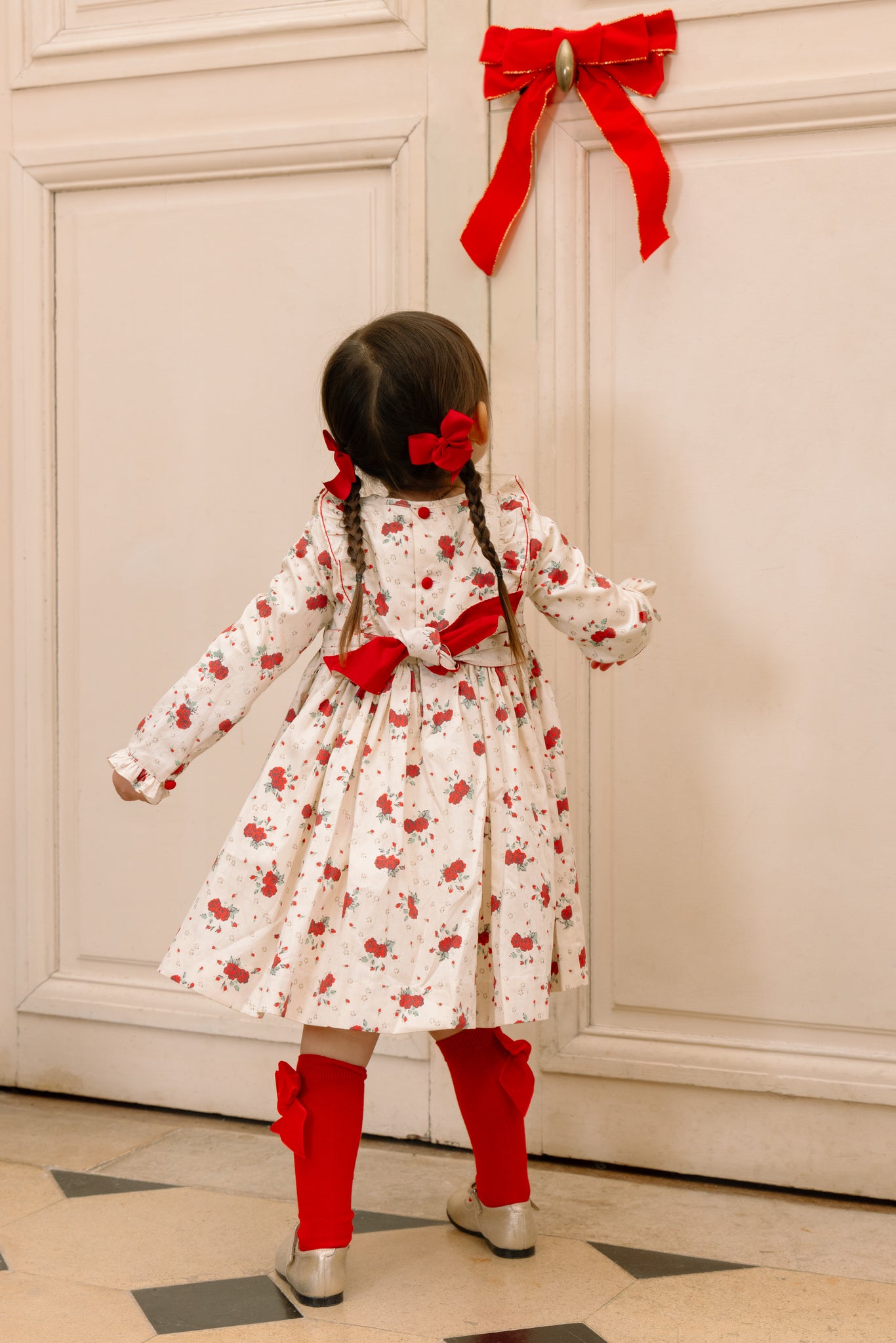 Child wearing Anastasia Christmas Smocked Dress and red boots standing in front of a door with a red bow.