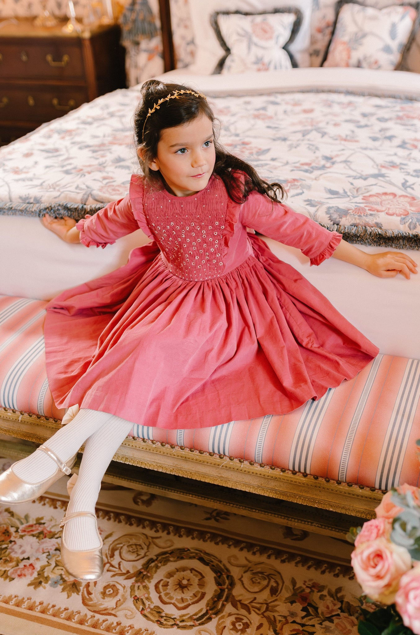 Girl in pink corduroy long-sleeve smocked dress with flutter sleeves and reversible bow, sitting in a Parisian room.