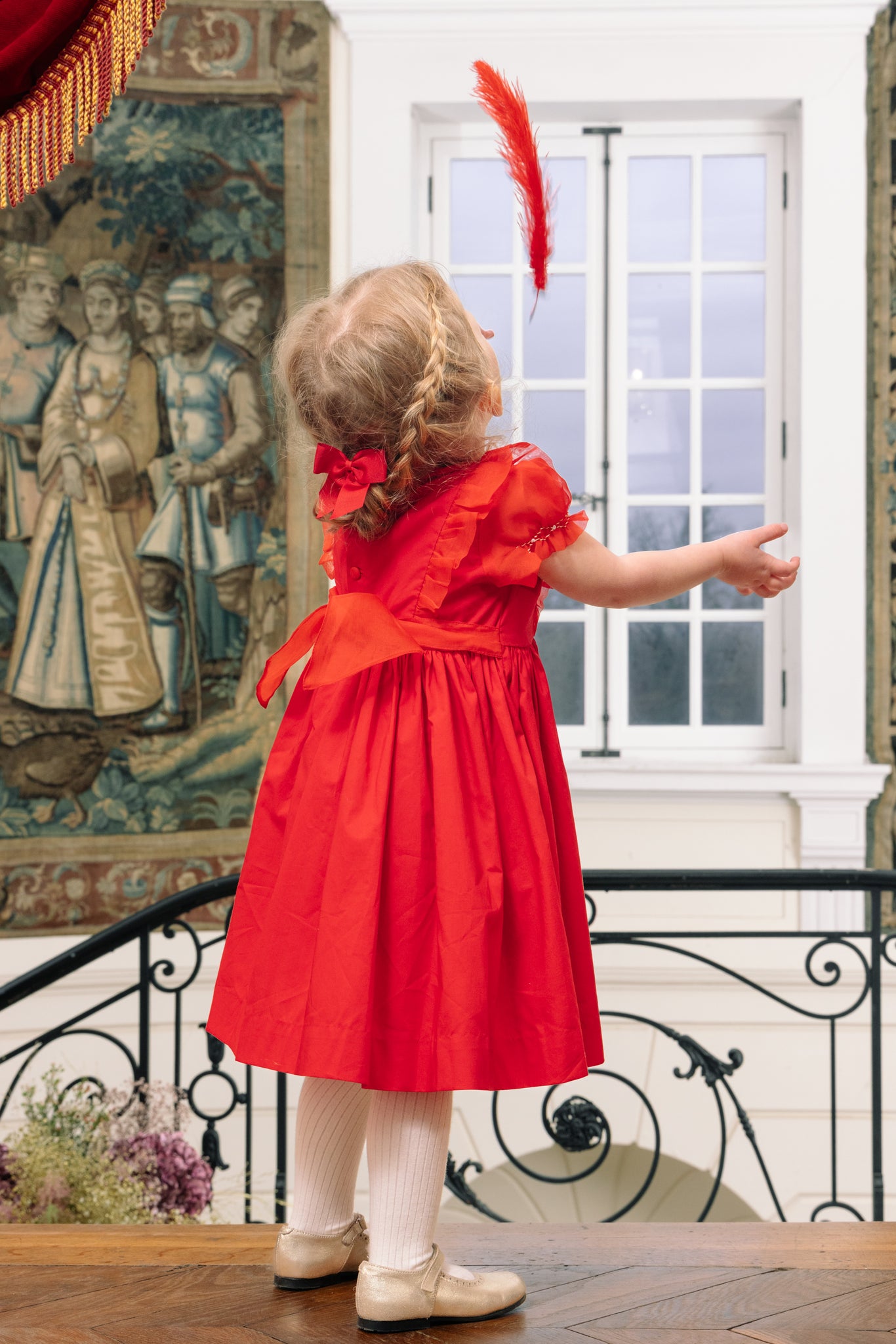 Child in Camelia Red Cotton Smocked Dress standing in a room with decorative elements
