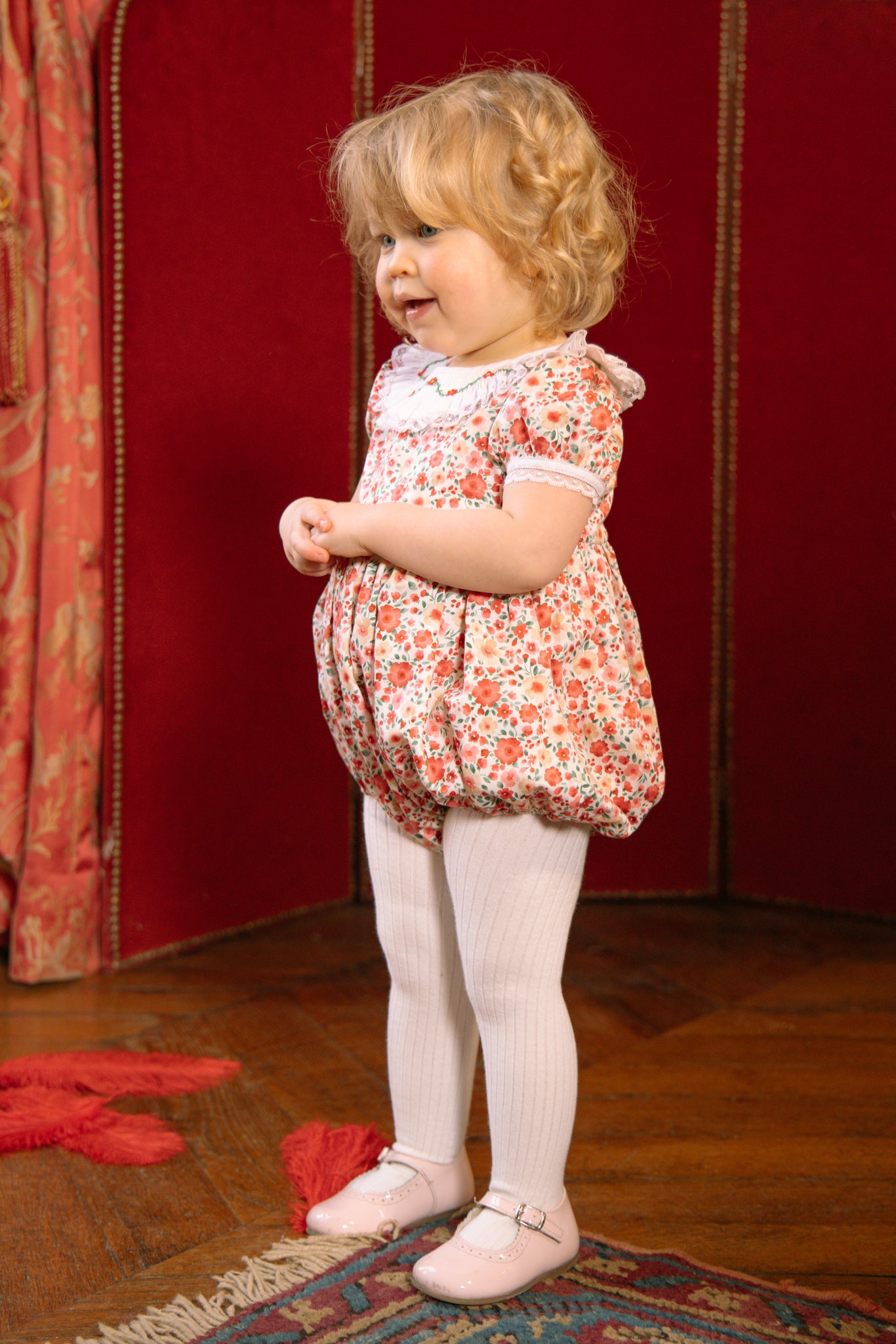 Young child in Celestine Red Floral Smocked Romper standing on a wooden floor with red curtains in the background