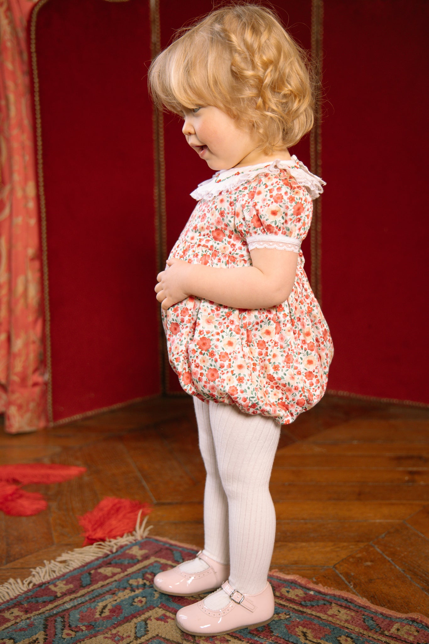 Young child in Celestine Red Floral Smocked Romper standing on a wooden floor with a red wall in the background