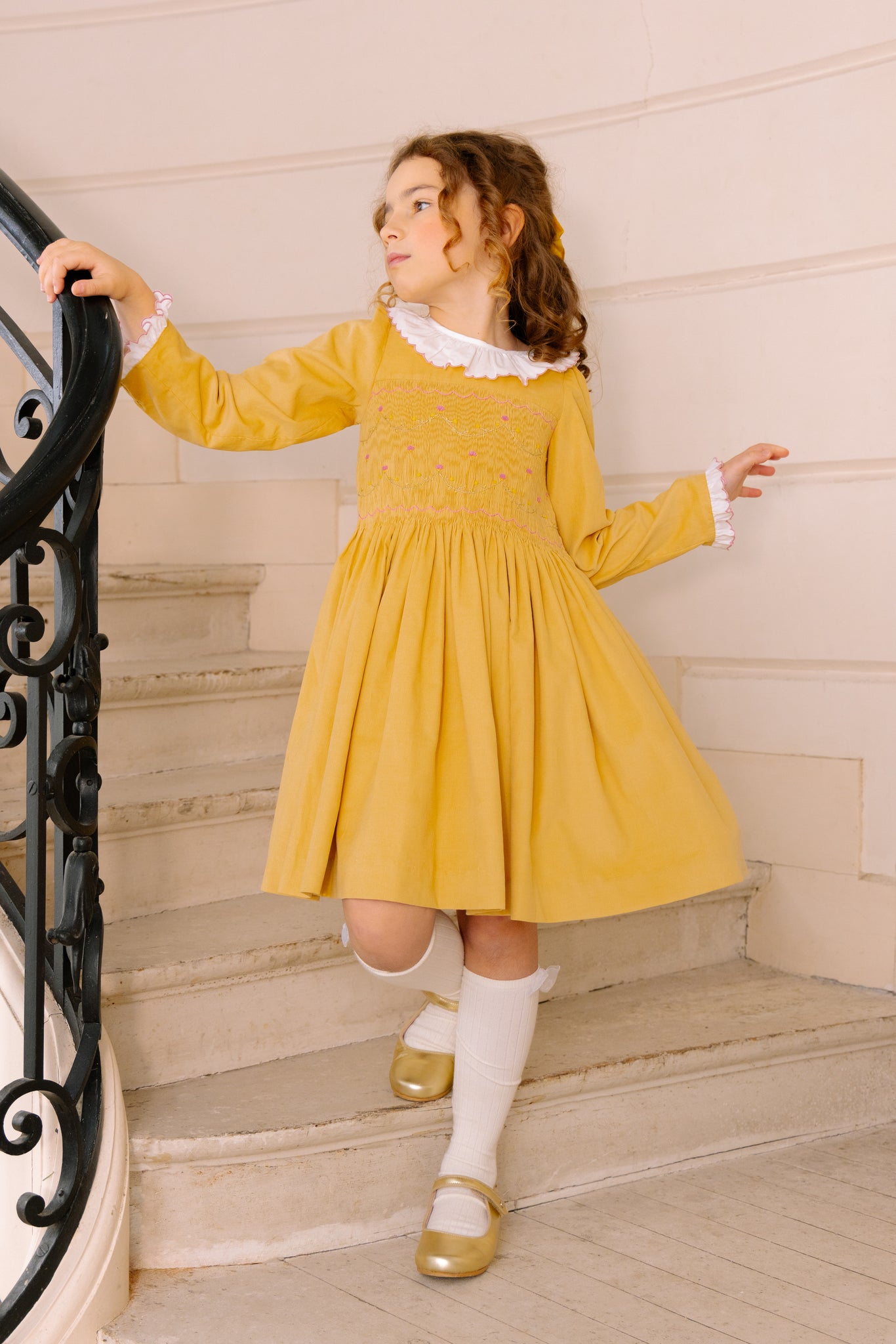 A young girl wearing Chiara Yellow Smocked Dress, paired with white socks and gold shoes, standing on a staircase.