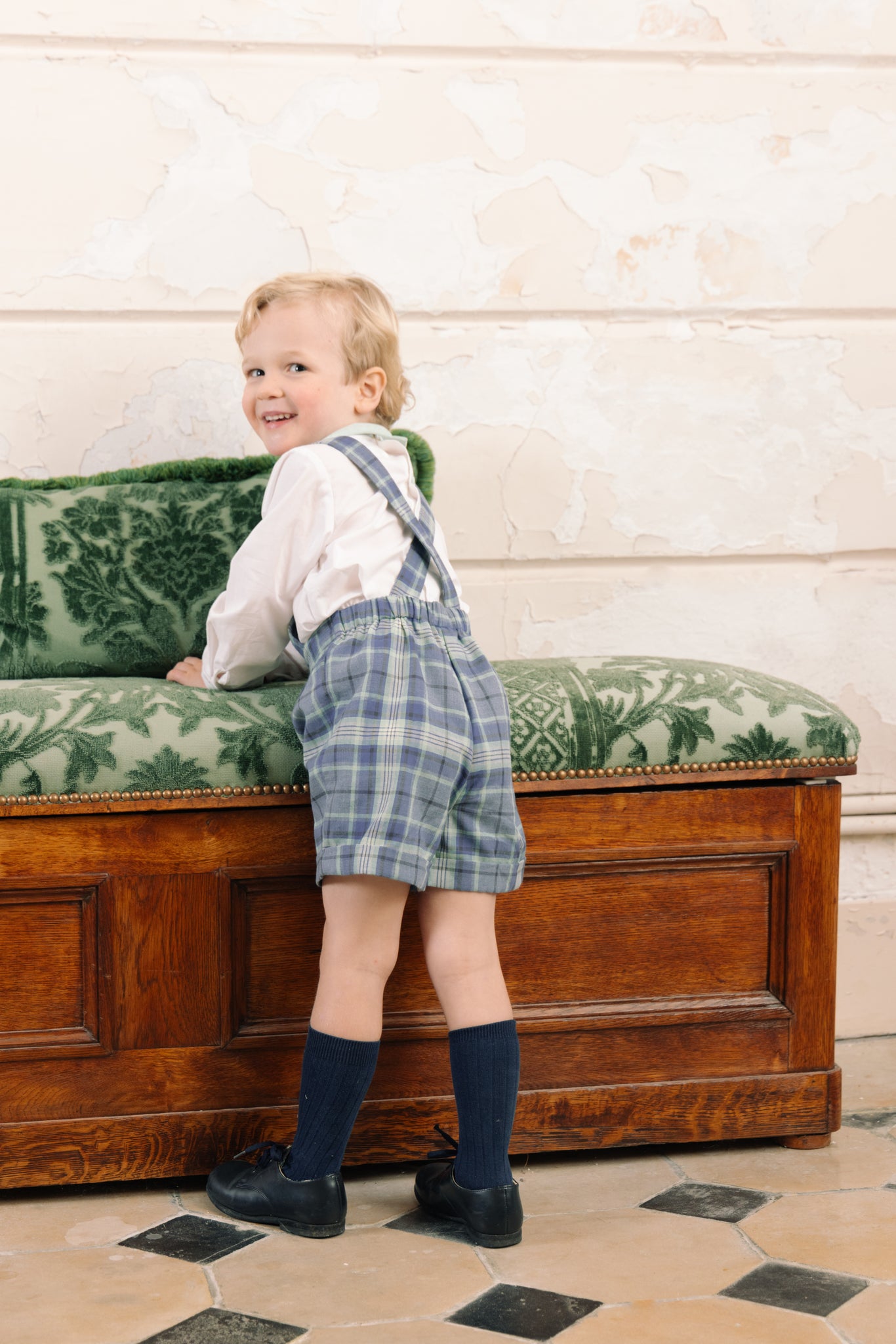 Child in George Blue Tartan Overalls and Shirt standing on a wooden bench with green cushion against a white wall.