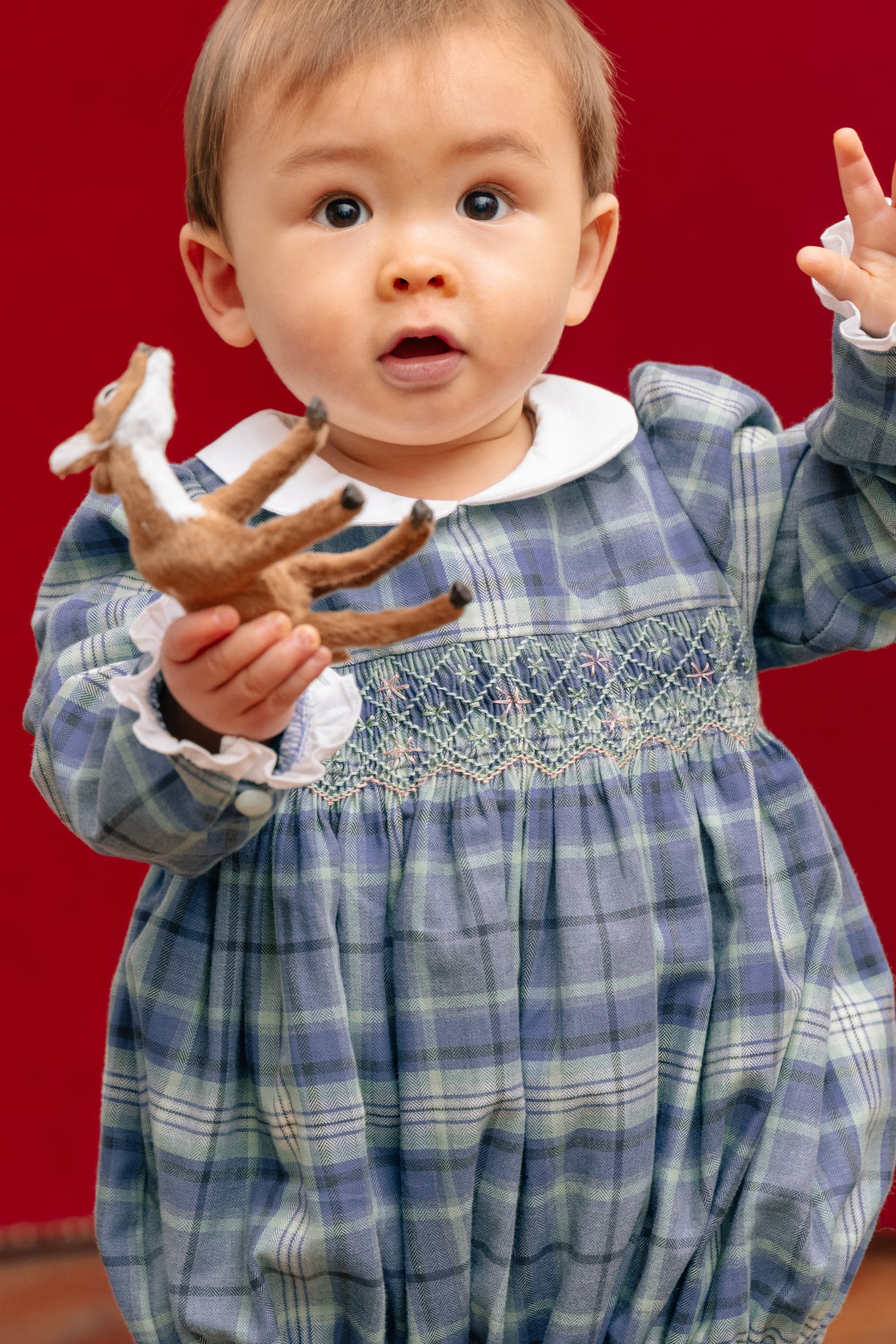 Child in Heidi Blue Tartan Baby Romper holding a toy against a red background