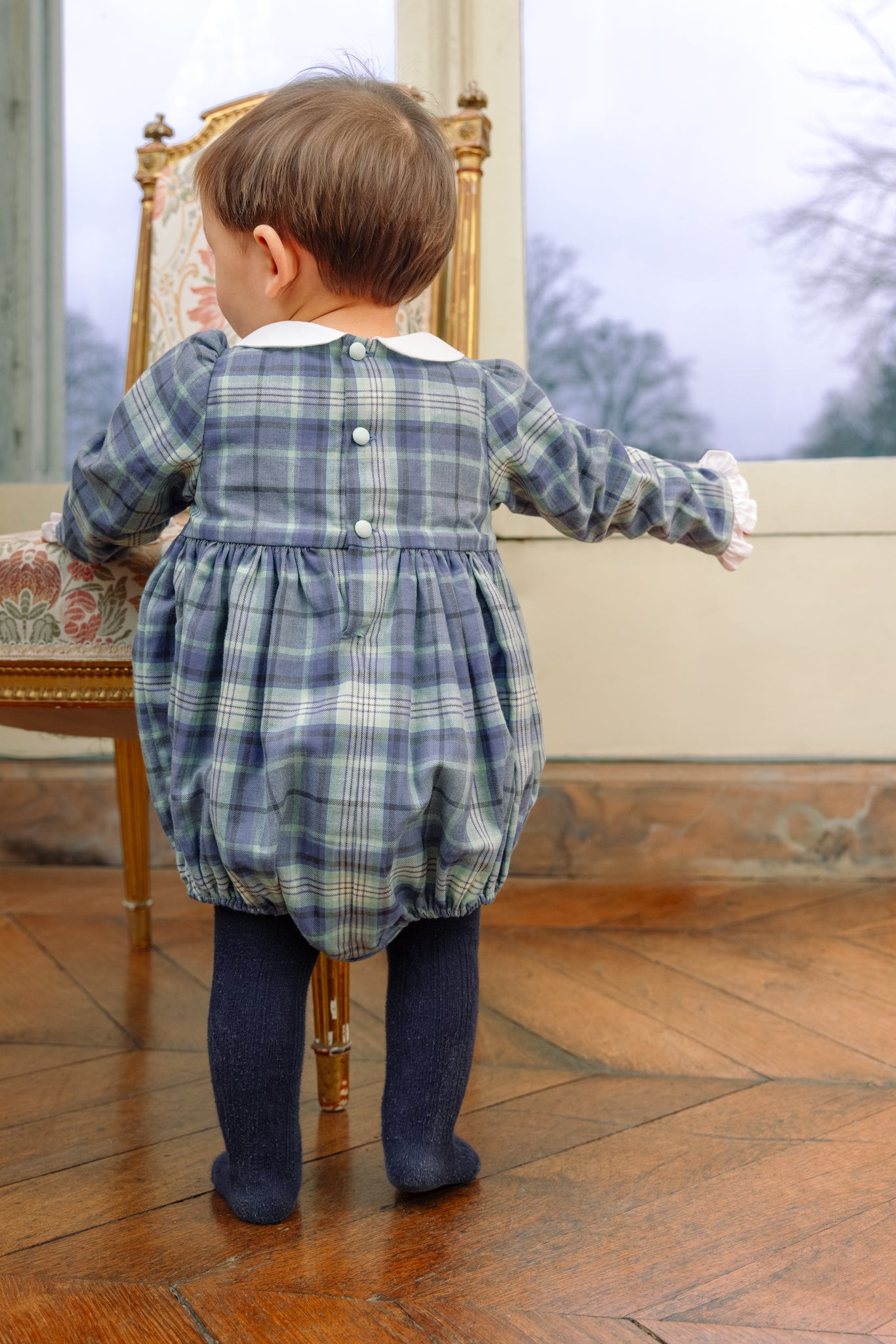 Child wearing Heidi Blue Tartan Baby Romper standing on a wooden floor.