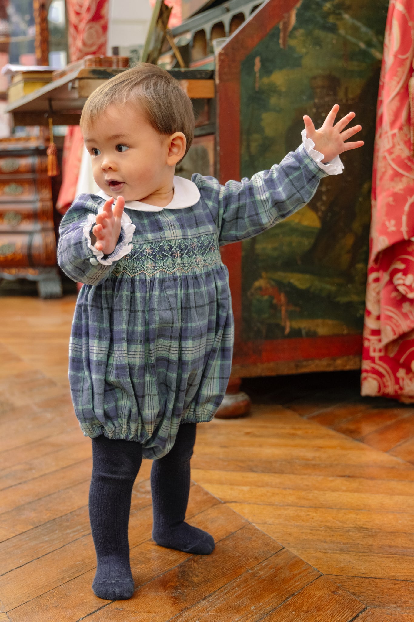 Baby in blue tartan smocked romper with white collar, green-and-white smocking, long sleeves, and elastic leg openings.