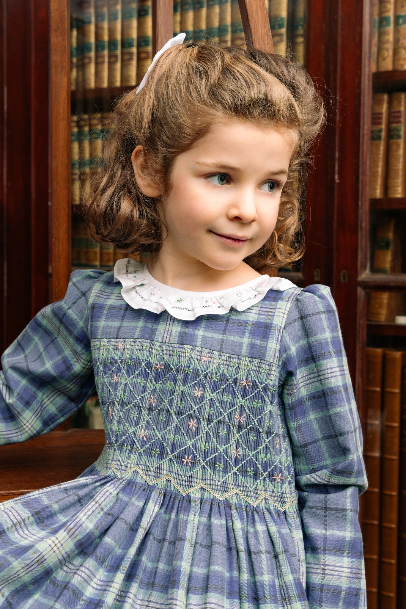 Young girl in Heidi Blue Tartan Smocked Dress standing in front of bookshelves.