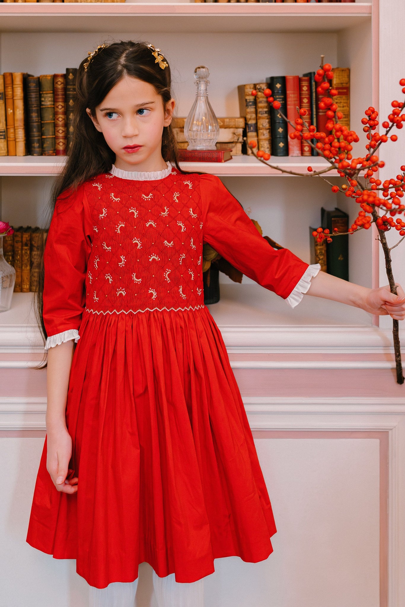 Girl in red long-sleeve smocked dress with white-gold moon embroidery, frilled collar, and cuff ruffles, posing in a Parisian room.