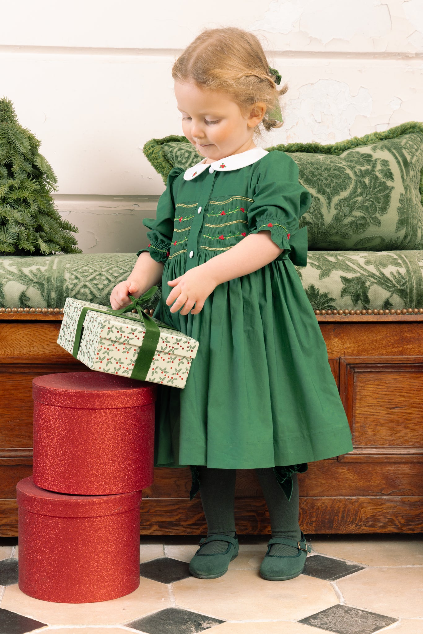 Young girl in Olympe Green Smocked Christmas Dress opening a gift box in a decorated room.