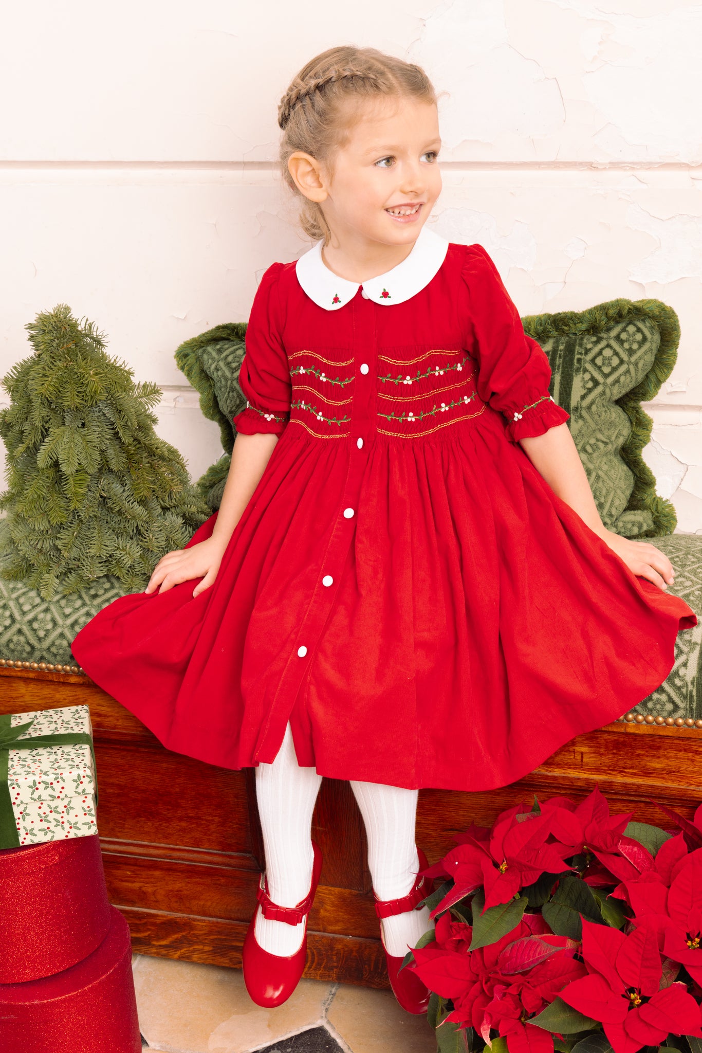 Young girl in Olympe Red Holiday Smocked Dress, sitting on a wooden bench with Christmas decorations.
