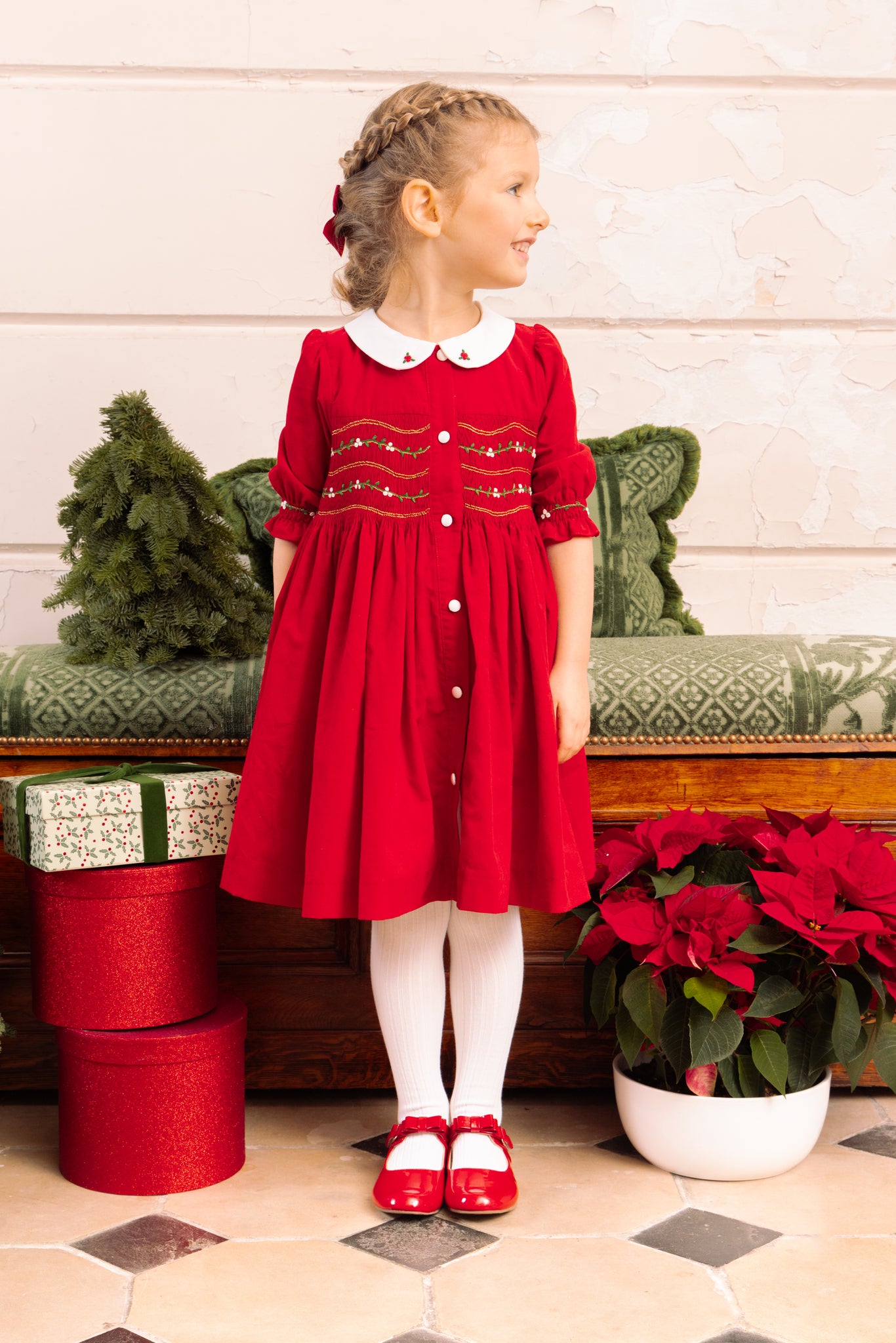 Girl in red smocked dress with Peter Pan collar and holly embroidery, ¾ sleeves with embroidered cuffs, wearing matching bloomers.