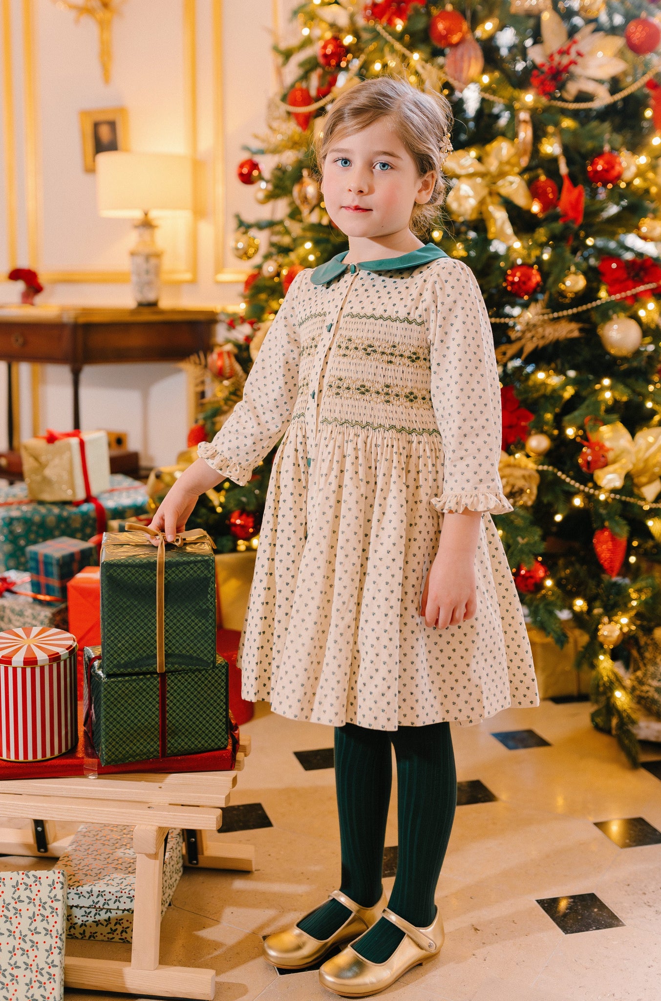 Girl in green long-sleeve smocked dress with Peter Pan collar, French holiday print, and reversible bow, standing by a decorated Christmas tree.