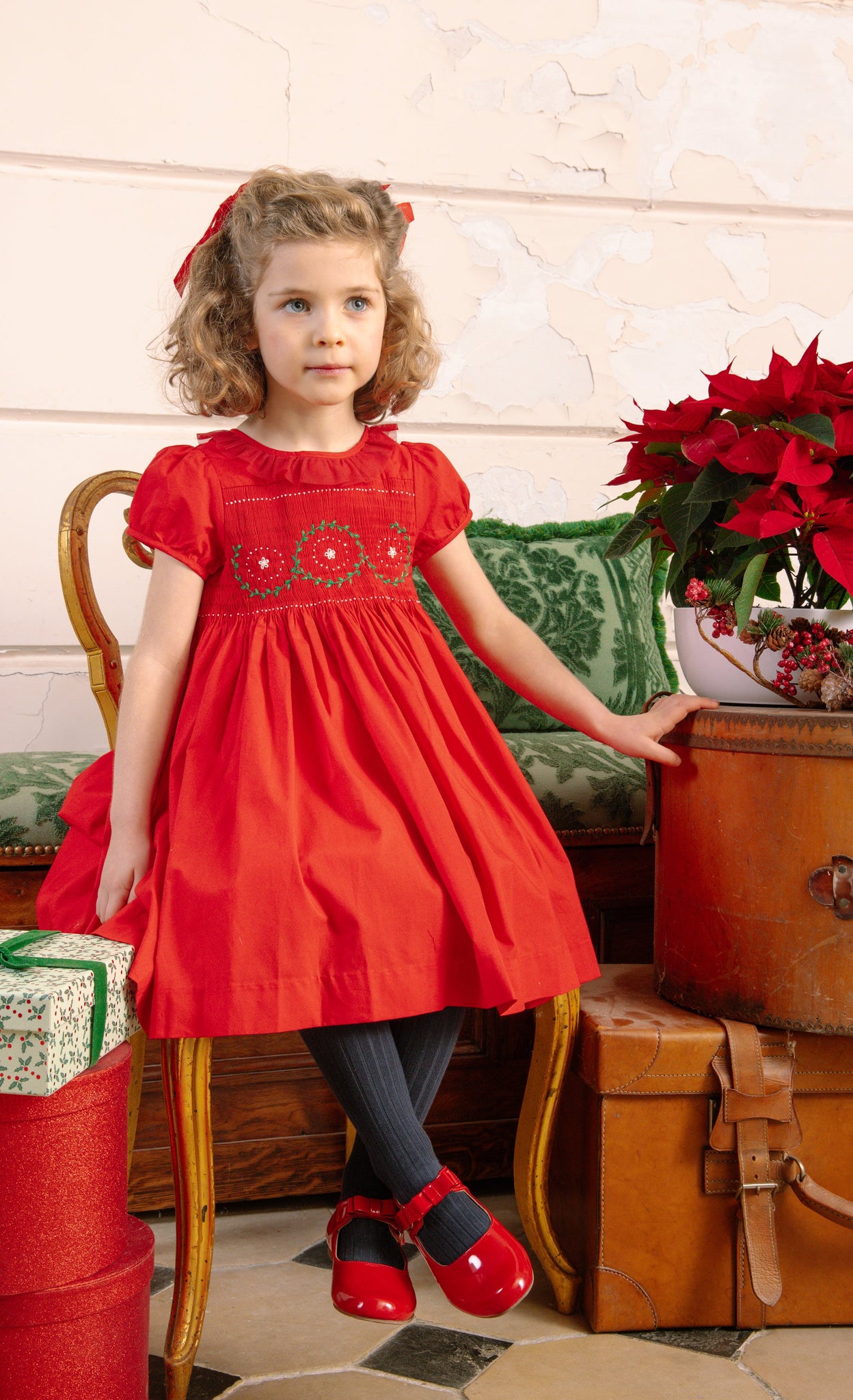 Young girl in Sofia Red Smocked Dress standing next to vintage suitcases and a potted plant.