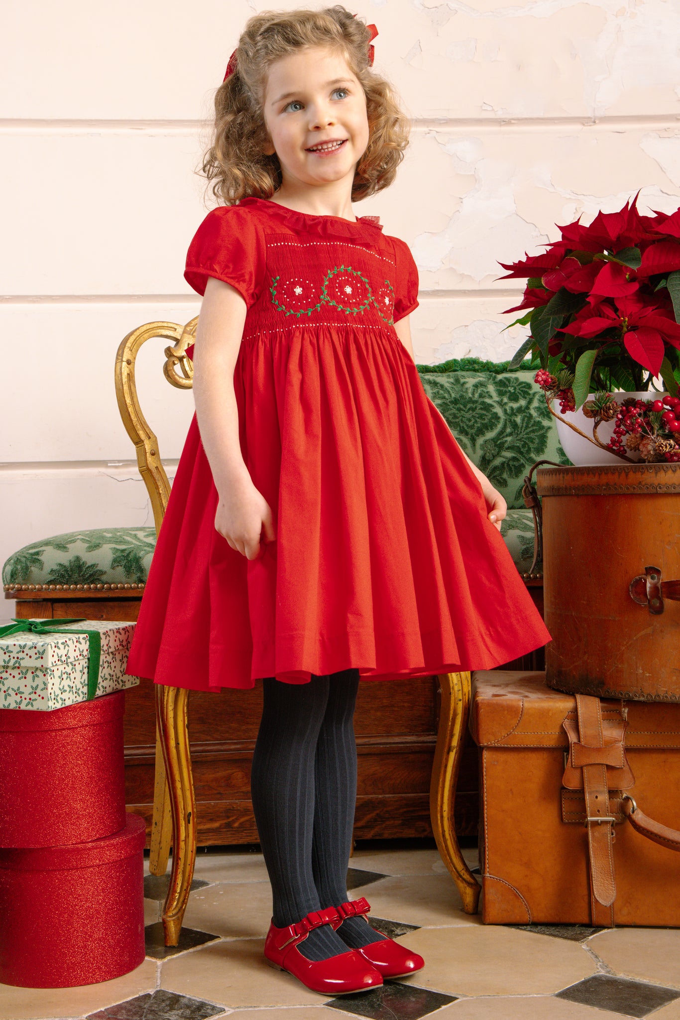 Young girl in Sofia Red Smocked Dress standing in a room with decorative items.