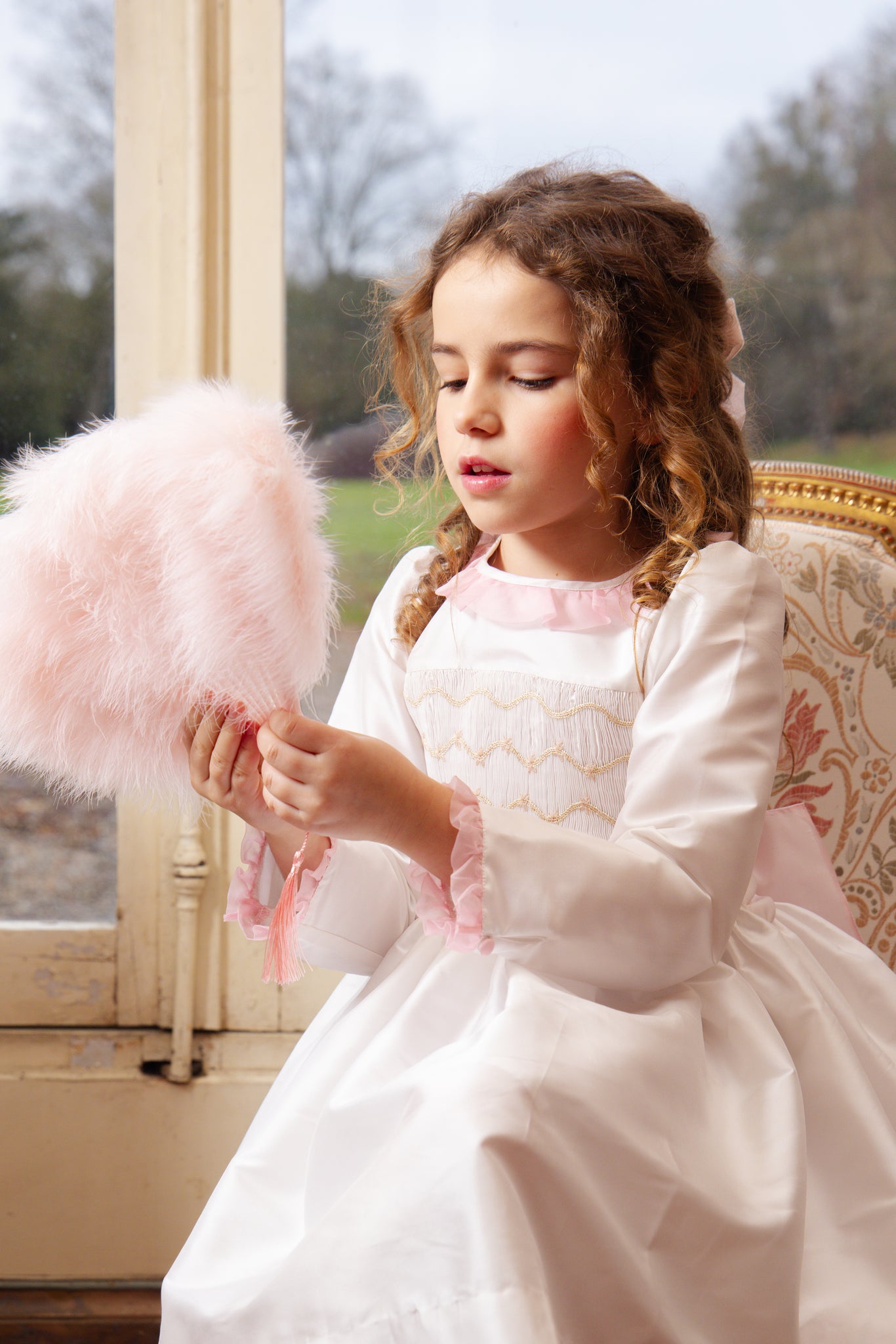Young girl in Sofia White Taffeta Long Sleeve Smocked Dress holding a pink fluffy object indoors.