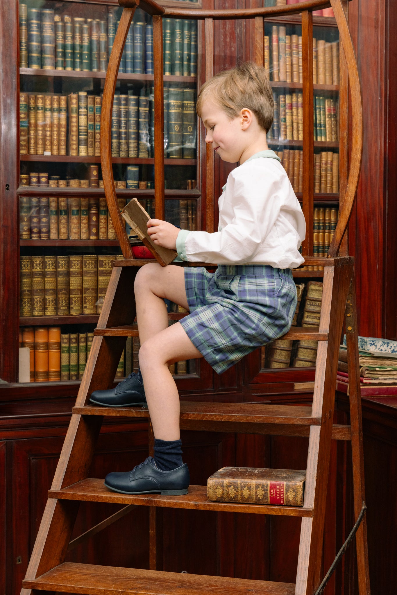 Child sitting on a wooden ladder in a library surrounded by books.