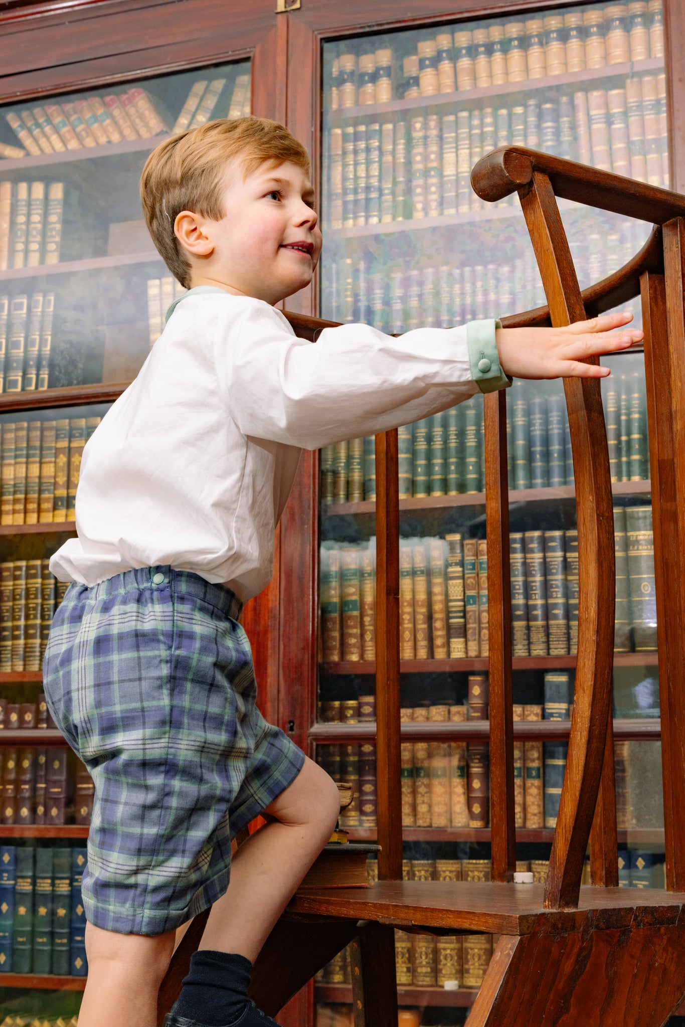 Young boy standing on a chair in front of a bookshelf filled with books.